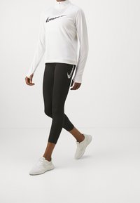 Maglietta bianca a maniche lunghe con zip a metà e logo Nike nero, abbinata a leggings neri con logo Nike bianco. Le scarpe sono sneaker bianche.