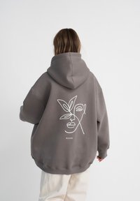 Felpa grigia oversize con cappuccio regolabile. Presenta un design in tracciato bianco di un volto e una foglia sul retro, con orlo a costine.
