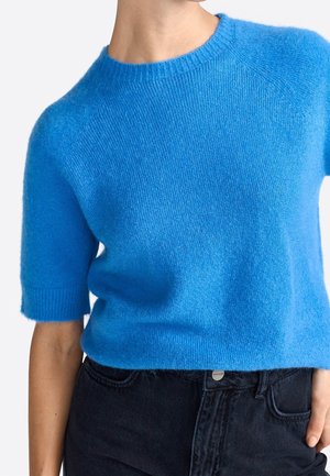 Lyseblå kortærmet strikket sweater båret med sort højtaljede jeans, som viser overkroppen og venstre arm.