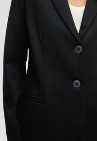 Zwarte blazer met twee knopen dichtgeknoopt, gedragen over een witte ribbeltop, die het bovenlichaam en een deel van de linkerarm toont.