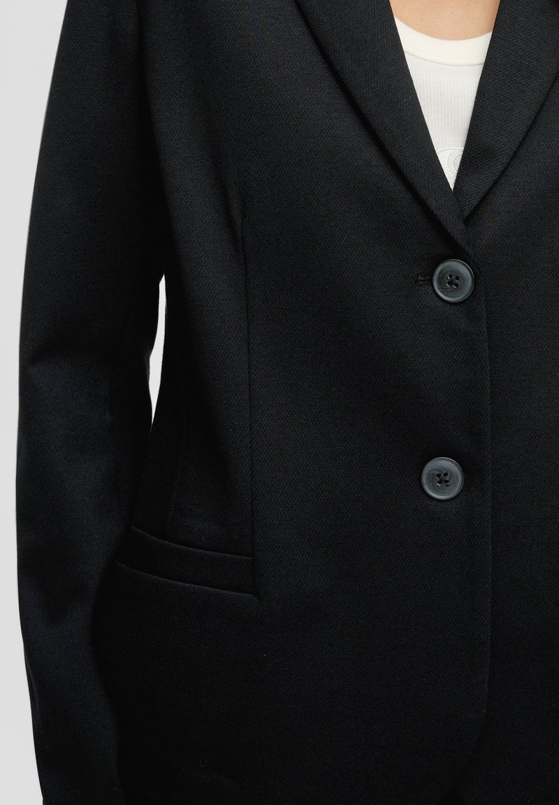 Blazer noir avec deux boutons fermés, porté par-dessus un haut à côtes blanc, montrant le haut du torse et une partie du bras gauche.