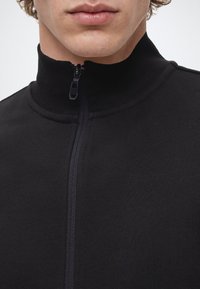 Gros plan d'une personne portant une veste noire zippée jusqu'au cou, montrant uniquement les lèvres et le menton.