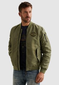 Homme portant une veste bomber vert olive avec des écussons et un jean bleu, regardant sur le côté sur un fond uni.