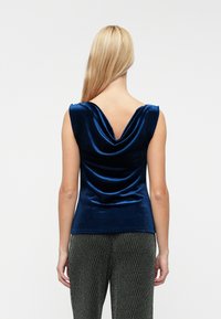 Cowl-back, sleeveless top in deep blue velvet, featuring a draped design and a fitted silhouette, paired with textured gray patterned trousers.