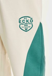 Hvit hettegenser med grønne detaljer, med brodert FC ECKŌ UNLIMITED-logo og et grafisk design med trekantede former.