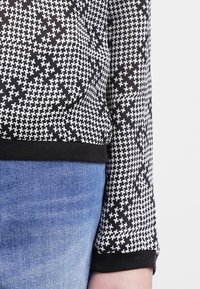 Haut noir et blanc à motif pied-de-poule, avec des manches longues et un ourlet côtelé noir. Porté sur un jean en denim bleu.