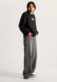 Sweat-shirt noir avec un patch logo blanc, associé à un jean large gris et des chaussures slip-on à damier noir et blanc.