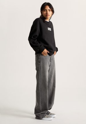 Sweat-shirt noir avec un patch logo blanc, associé à un jean large gris et des chaussures slip-on à damier noir et blanc.