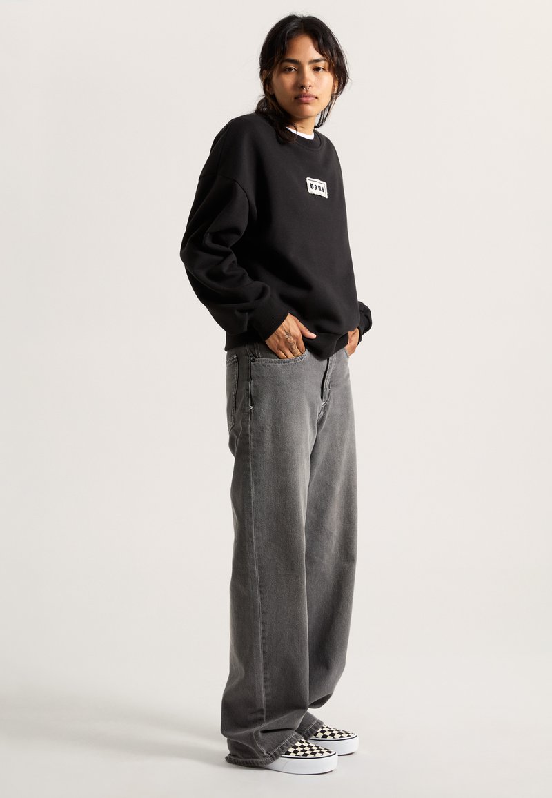 Sweat-shirt noir avec un patch logo blanc, associé à un jean large gris et des chaussures slip-on à damier noir et blanc.