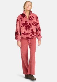 Fleece-Pullover in Pink mit burgunderfarbenem Blättermuster; mit einem halben Reißverschluss am Kragen und elastischen Bündchen ausgestattet. Kombiniert mit rosa Cordhosen und dunklen Stiefeln.
