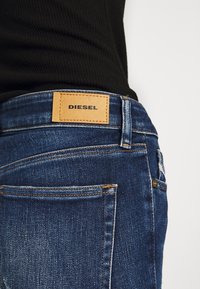 Jeans de mezclilla en azul oscuro con un parche de cuero etiquetado como "DIESEL" en la cintura. Presentan costuras en contraste y múltiples bolsillos traseros.