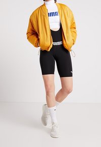 Żółta siatkowa kurtka bomberka nałożona na biało-niebieską koszulkę z logo, połączona z czarnymi szortami i białymi skarpetkami, dopełniona jasnymi sneakersami.
