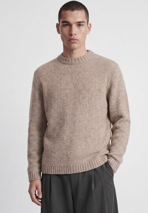 Jeune homme aux cheveux courts portant un pull beige en maille et un pantalon foncé, debout devant un fond clair uni.