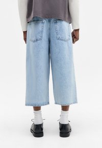 Pantalones culottes de denim azul claro con un corte holgado, presentando dos bolsillos traseros y un dobladillo sin terminar. Combinados con zapatos negros y calcetines blancos.