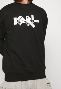 Czarna bluza z białym geometrycznym logo „KARL” na przodzie. Posiada okrągły dekolt i ściągacze przy mankietach dla dopasowanego wyglądu.