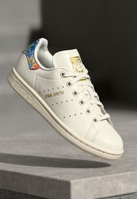 Valge nahast Adidas Stan Smith jalgpallijalats kuldse logoga, värvilise lillemustriga aksent konts ning tekstureeritud kummitald.