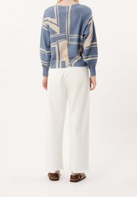 Personne portant un pull à motifs géométriques bleu et beige, un pantalon large blanc à ourlets bruts, et des sandales marron à enfiler avec des boucles, debout.