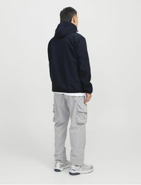 Marineblauwe hoodie-jack met een gladde textuur, gecombineerd met lichtgrijze cargobroek met zijzakken. Model draagt zilveren sneakers.