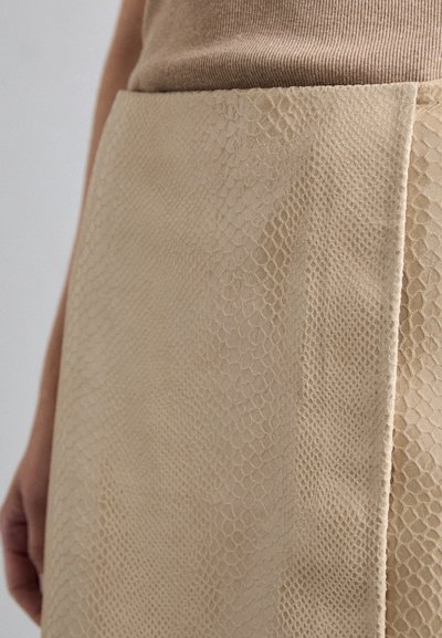 Beige szoknya domború anyagból, amely kígyóbőrre emlékeztet, sima felülettel és finom pikkelymintázattal.