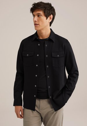 Sort skjorte med knapper lavet af glat stof, med to brystlommer, slim fit og krave. Bæres over en sort T-shirt.