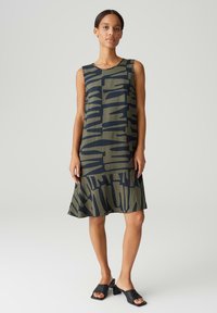 Sleeveless dress in olive green with bold navy abstract patterns. Flared hem with a textured finish, paired with black block-heeled sandals.