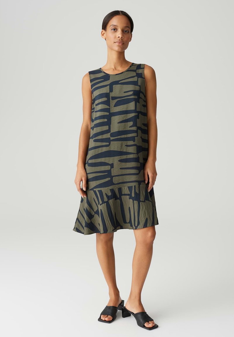 Sleeveless dress in olive green with bold navy abstract patterns. Flared hem with a textured finish, paired with black block-heeled sandals.