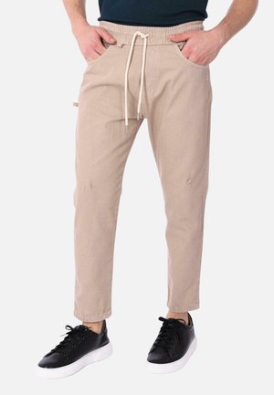 Pantaloni jogger beige con vita elasticizzata e cordoncino. Presentano due tasche laterali e dettagli strappati. Indossati con sneaker nere.