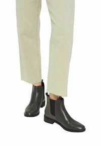 Harmaat nahkasaappaat nilkkapituisilla, joissa on sileä pinta, joustavat sivupaneelit ja teksturoitua yksityiskohtaa. Yhdistetty vaaleisiin, repaleiseen lahkeeseen.