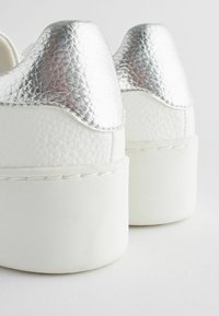Białe sneakersy na platformie z teksturowanego białego materiału i błyszczącymi srebrnymi akcentami z tyłu. Okrągły fason pięty, minimalne detale przeszyć.