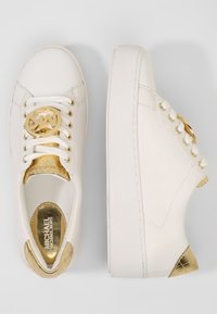 Sneakers bianchi con una texture liscia, logo dorato e dettagli. Presentano occhielli tono oro e un tallone metallico con motivo. Design con punta rotonda.