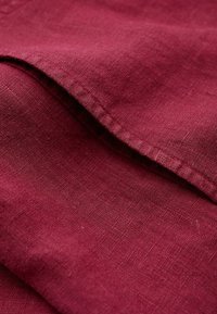 Burgundylinne med en slät textur och en diskret vävstruktur. Kanterna är snyggt sytt med kontrasterande sömmar.