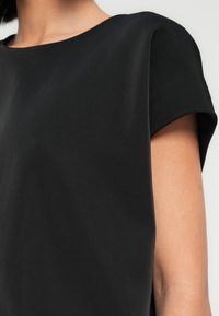 Μαύρο υφασμάτινο τοπ με κοντά, γυρισμένα μανίκια, στρογγυλή λαιμόκοψη, λείο ύφασμα και εφαρμοστή γραμμή κοντά στο σώμα.