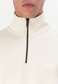 Light beige half-zip pullover made from soft fabric, featuring a black zipper and a square white logo on the chest. Smooth texture.