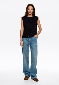Czarny top bez rękawów z rozrzuconymi srebrnymi ozdobami, zestawiony z jasnoniebieskimi szerokimi dżinsami i brązowym obuwiem. Gładka tkanina i luźny krój.