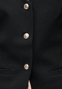 Veste noire sur mesure avec trois boutons métalliques dorés gravés d'un logo "G", avec des poches fendues de chaque côté.