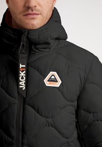 Svart vadderad jacka med hög krage, kviltad textur och en vit hexagonal logo med oranga accenter på vänster bröst. Dragkedjedetaljer synliga.