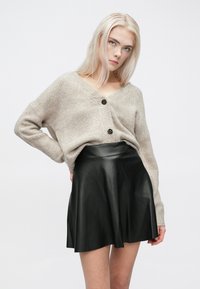 Beige kötött kardigán V-nyakkal és fekete gombokkal, fekete bőrből készült, bő szoknyával párosítva. Simább textúrájú és laza szabású.