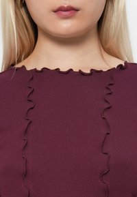 Burgundi színű top, bordázott textúrával, fodros dekoltárral a gallér mentén. Az anyag sima felületű és testhez simuló fazonú.