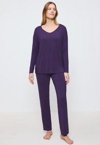 Top largo de manga larga morado con escote en V y abertura lateral, combinado con pantalones rectos morados a juego. Textura de tela suave y lisa.