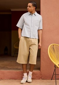 Jovem com cabelo trançado, vestindo uma camisa de manga curta com riscas, calças compridas bege, meias brancas e ténis, parado perto de uma parede rosa.