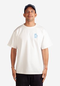 Bílý bavlněný tričko s krátkým rukávem, s modrou grafickou potiskem na levém prsu. Model má na sobě tmavé kordové kalhoty a námořnickou čepici.