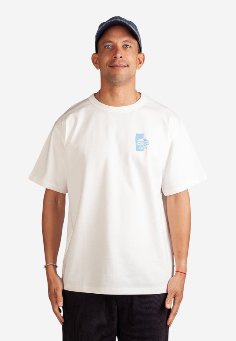 Bílý bavlněný tričko s krátkým rukávem, s modrou grafickou potiskem na levém prsu. Model má na sobě tmavé kordové kalhoty a námořnickou čepici.