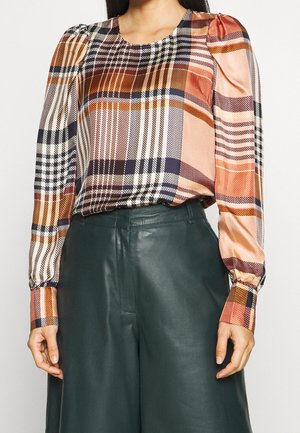 Femme portant un chemisier à manches longues à carreaux multicolores rentré dans un pantalon en cuir vert foncé taille haute, sur un fond uni.