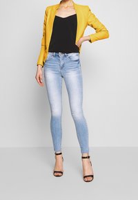 Blazer jaune sur un débardeur noir, associé à un jean skinny bleu clair et des talons noirs à lanières. Le tissu semble lisse ; un look décontracté mais soigné.