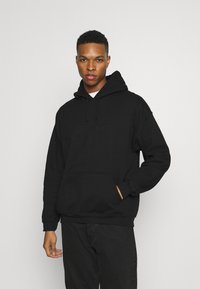 Sort bomulds-hoodie med en frontlomme, snørebånd i hætten og ribbede manchetter; båret med sorte bukser. En simpel og afslappet fit-design.