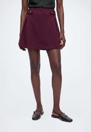 Femme portant une jupe courte bordeaux avec des détails de boutons et des chaussures plates noires avec des brides à la cheville, debout sur fond blanc.