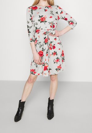 Kobieta w jasnoszarej sukience do kolan z czerwonym kwiatowym wzorem i rękawami do łokcia, czarne botki za kostkę, ręka na biodrze.