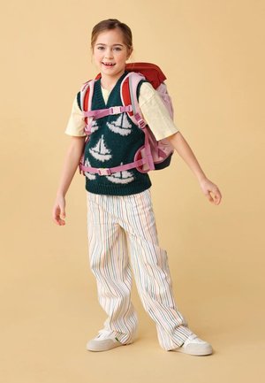 Ein Kind trägt einen grünen Pullover mit weißen Segelboot-Mustern, cremefarbene gestreifte Hosen und einen pinken und roten Rucksack. Weiße Turnschuhe runden das Outfit ab.