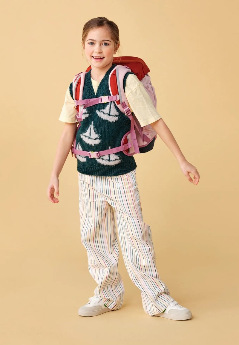 Bambino con un maglione verde decorato con modelli di velieri bianchi, pantaloni a righe crema e uno zaino rosa e rosso. Sneakers bianche completano l'outfit.