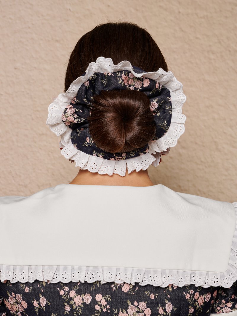 Γυναίκα με σκούρα μαλλιά δεμένα σε κότσο, στολισμένα με μεγάλο υφασμάτινο λαστιχάκι με λευκή δαντέλα, φοράει φλοράλ φόρεμα με λευκό γιακά.